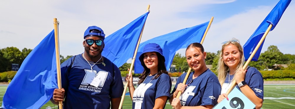 Four student orienation leaders smiling, holding blue flags and wearing blue and white Monmouth University apparrel