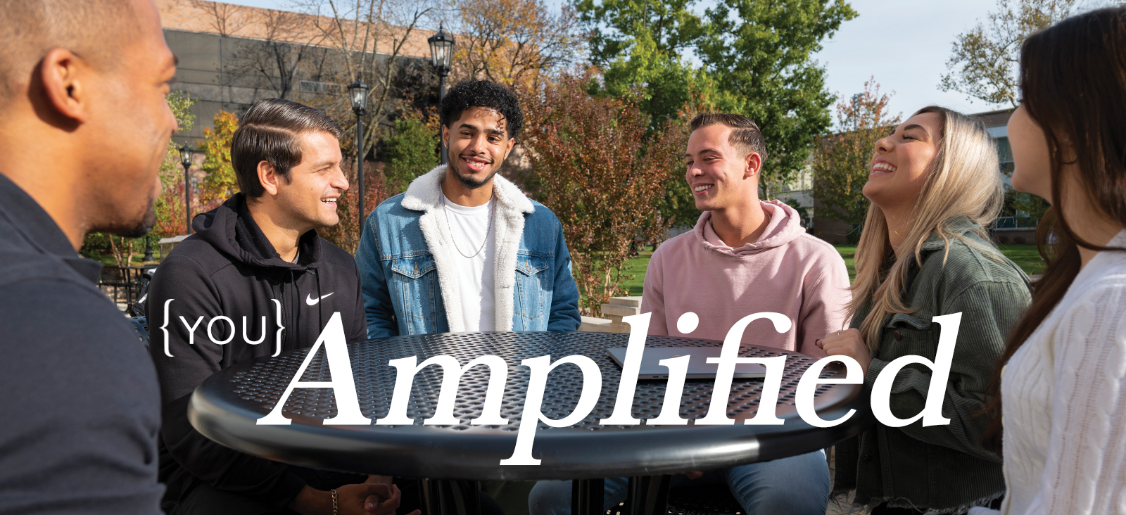 You, Amplified. Students smiling and talking at a table outside of a campus building
